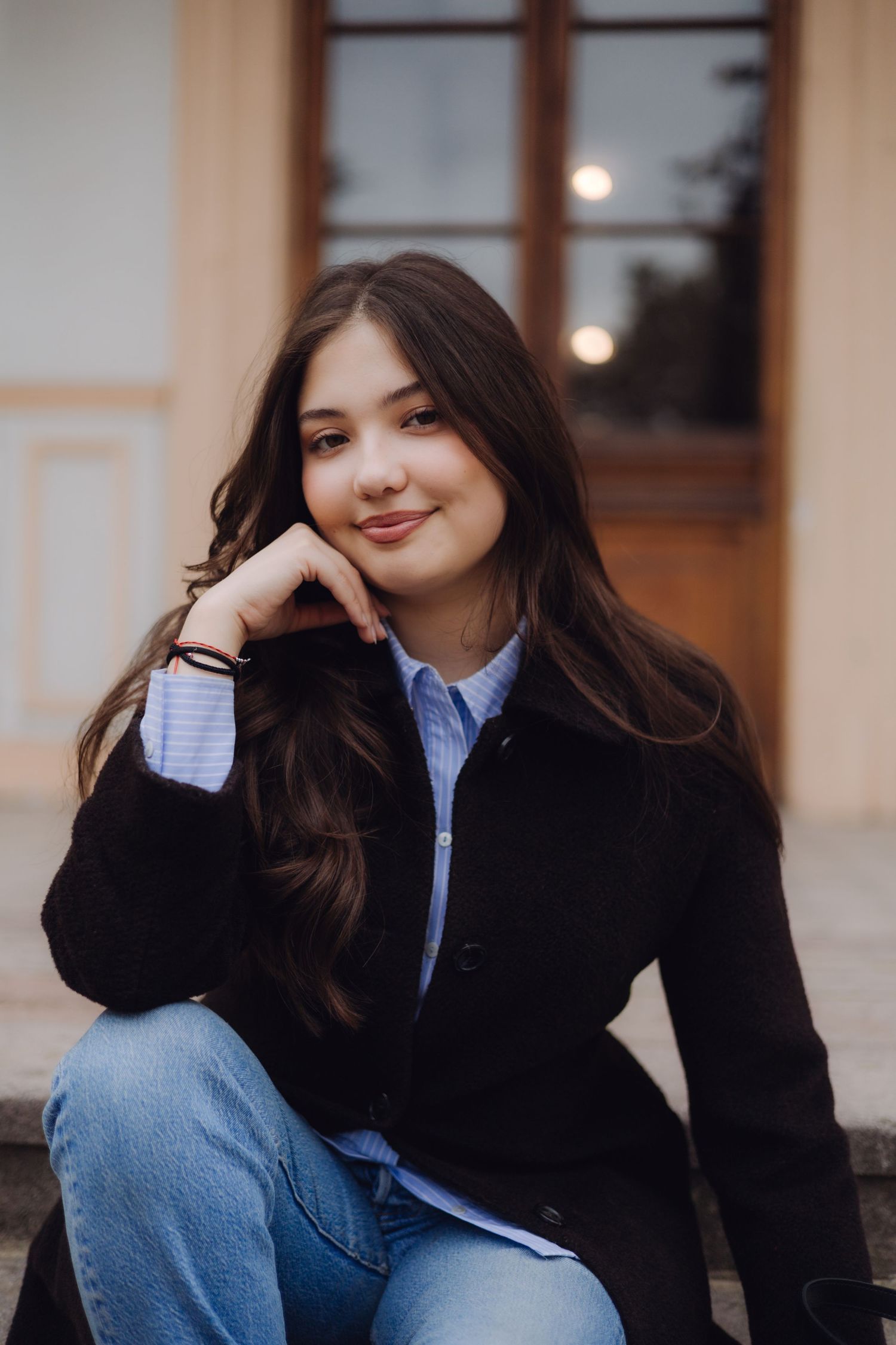 Young woman with long dark hair sits outdoors in black coat and jeans, resting chin on hand, smiling softly.