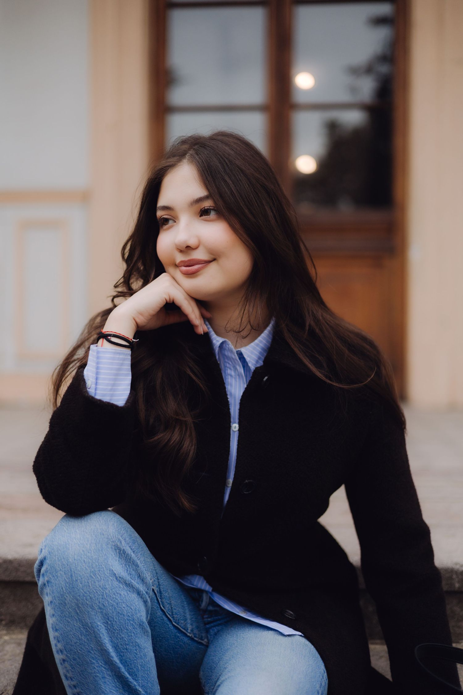 Young woman with long dark hair sits outdoors in black coat and jeans, smiling with hand on chin.