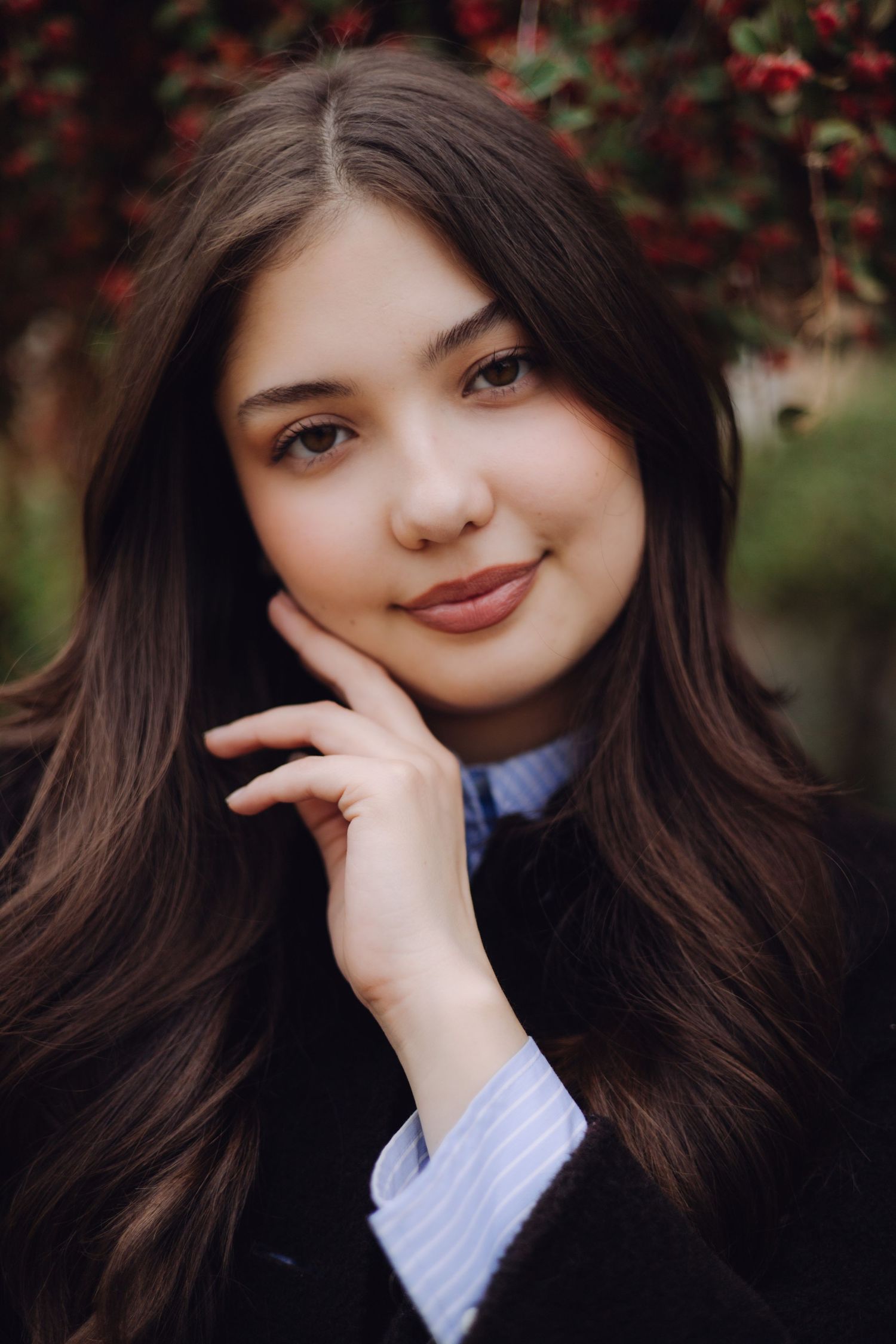Young woman with long dark hair smiling softly, resting chin on hand, wearing a blue shirt and dark coat outdoors.