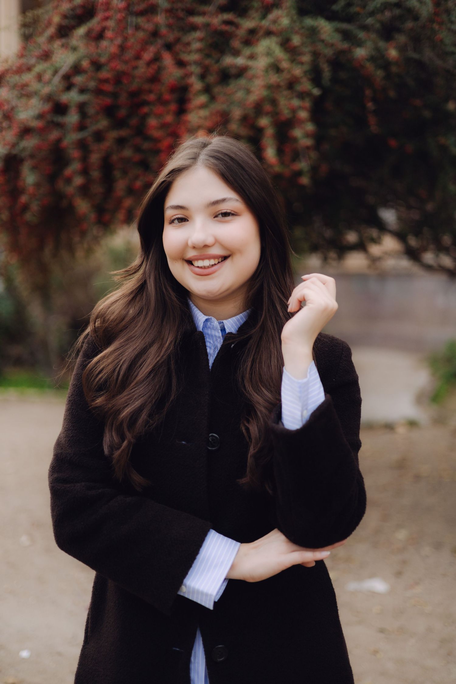 Smiling young woman with long dark hair wearing a black coat over a blue shirt outdoors.
