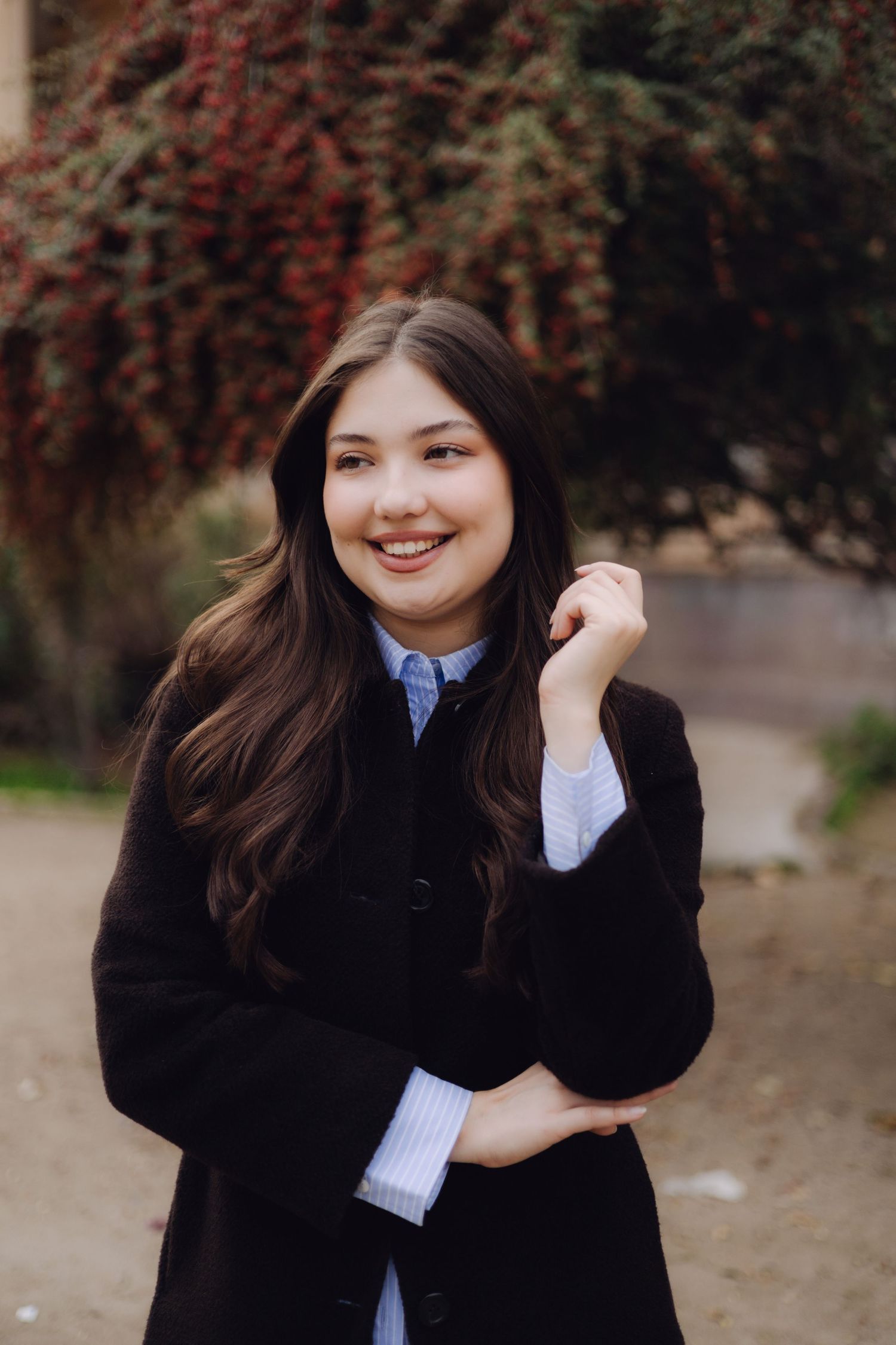 Young woman with long dark hair smiling outdoors in a black coat over a blue striped shirt, with red foliage behind her.