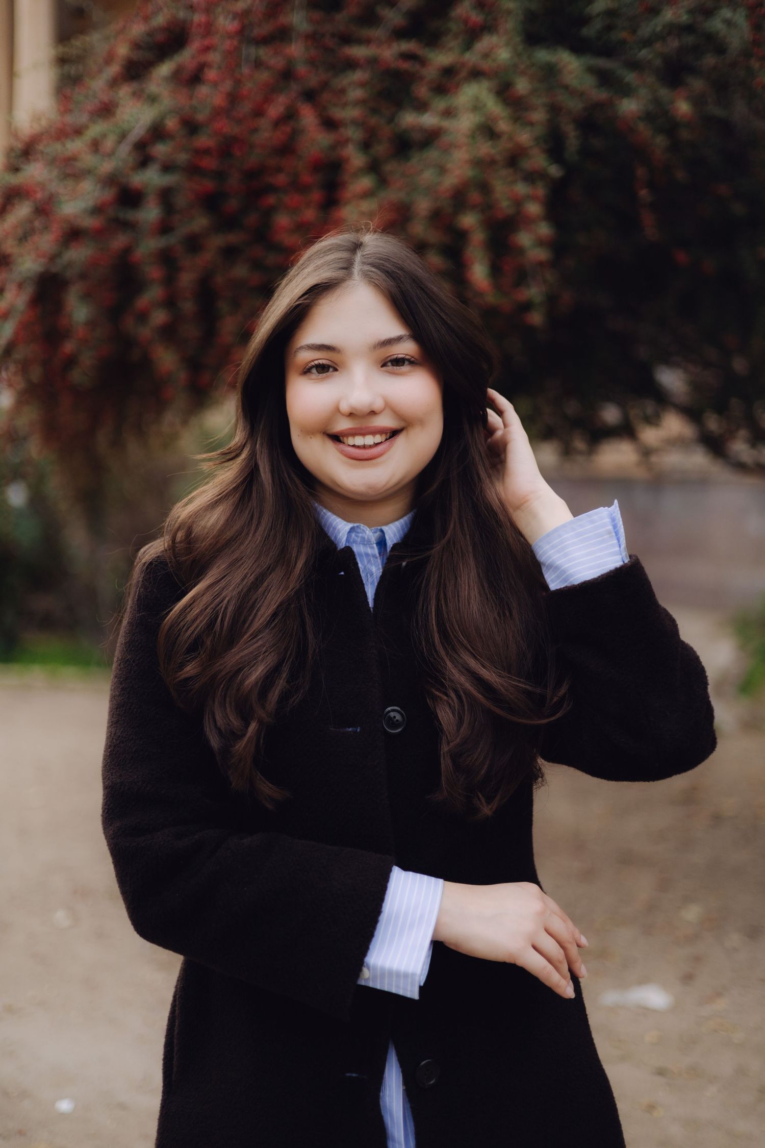 Young woman with long dark hair smiling outdoors, wearing a black coat over a blue striped shirt.