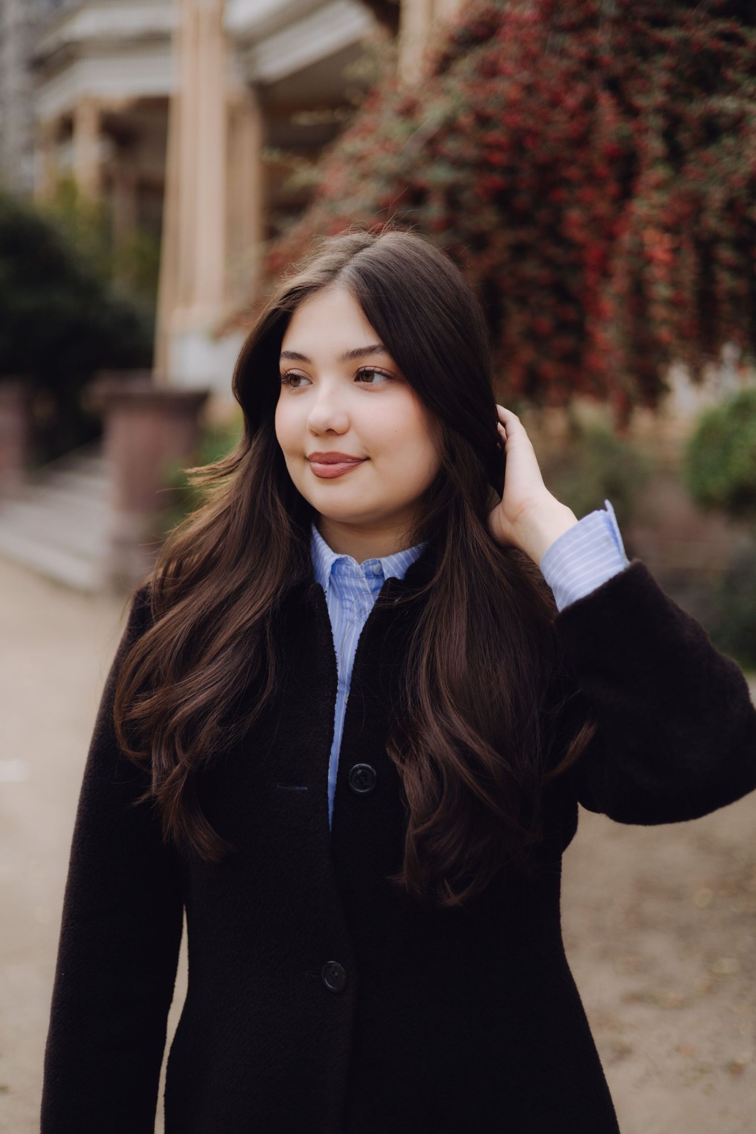 Young woman with long dark hair in a black coat posing outdoors near red foliage.