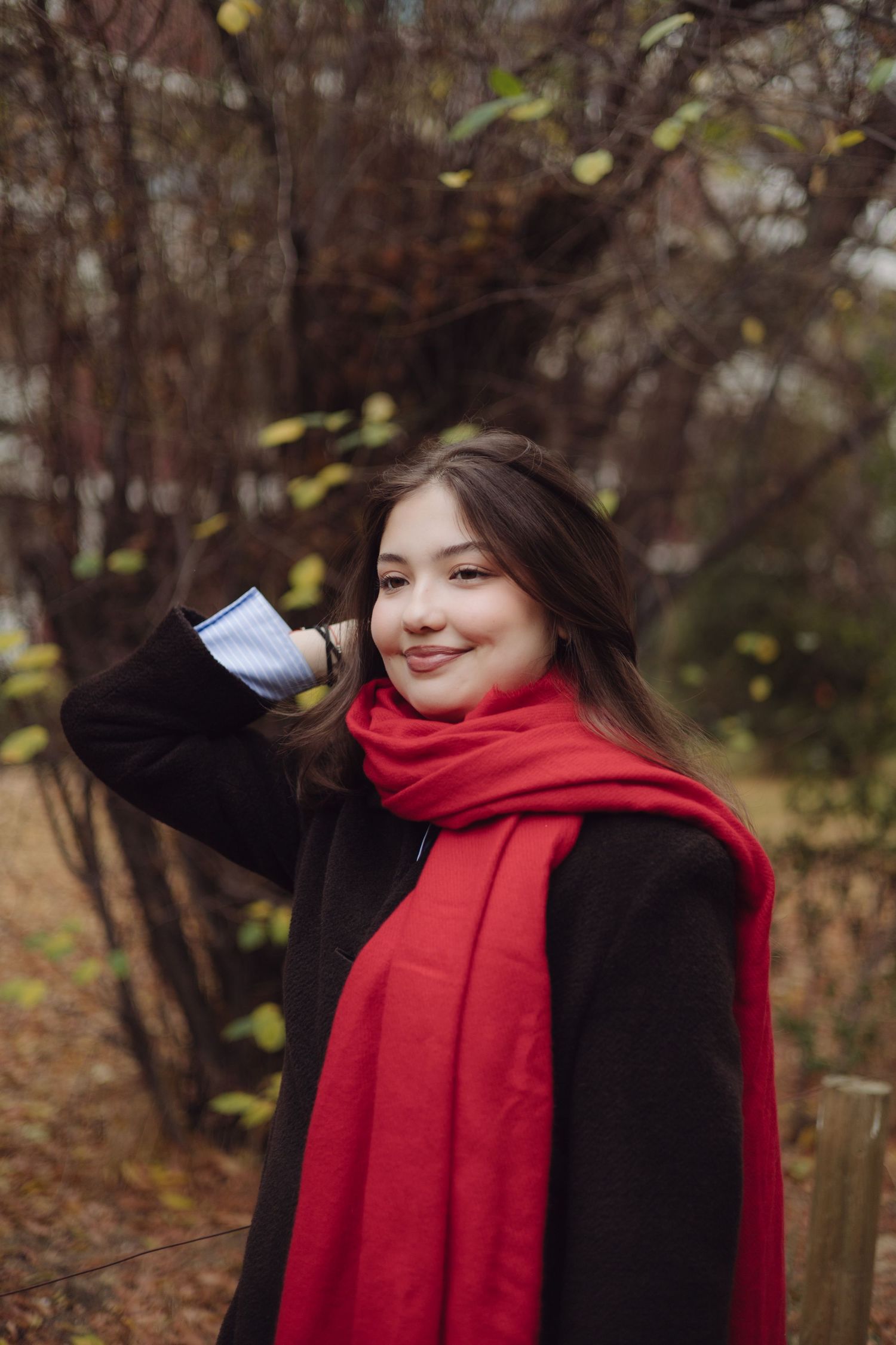Young woman with long brown hair wearing a red scarf and black coat smiling outdoors in autumn park.