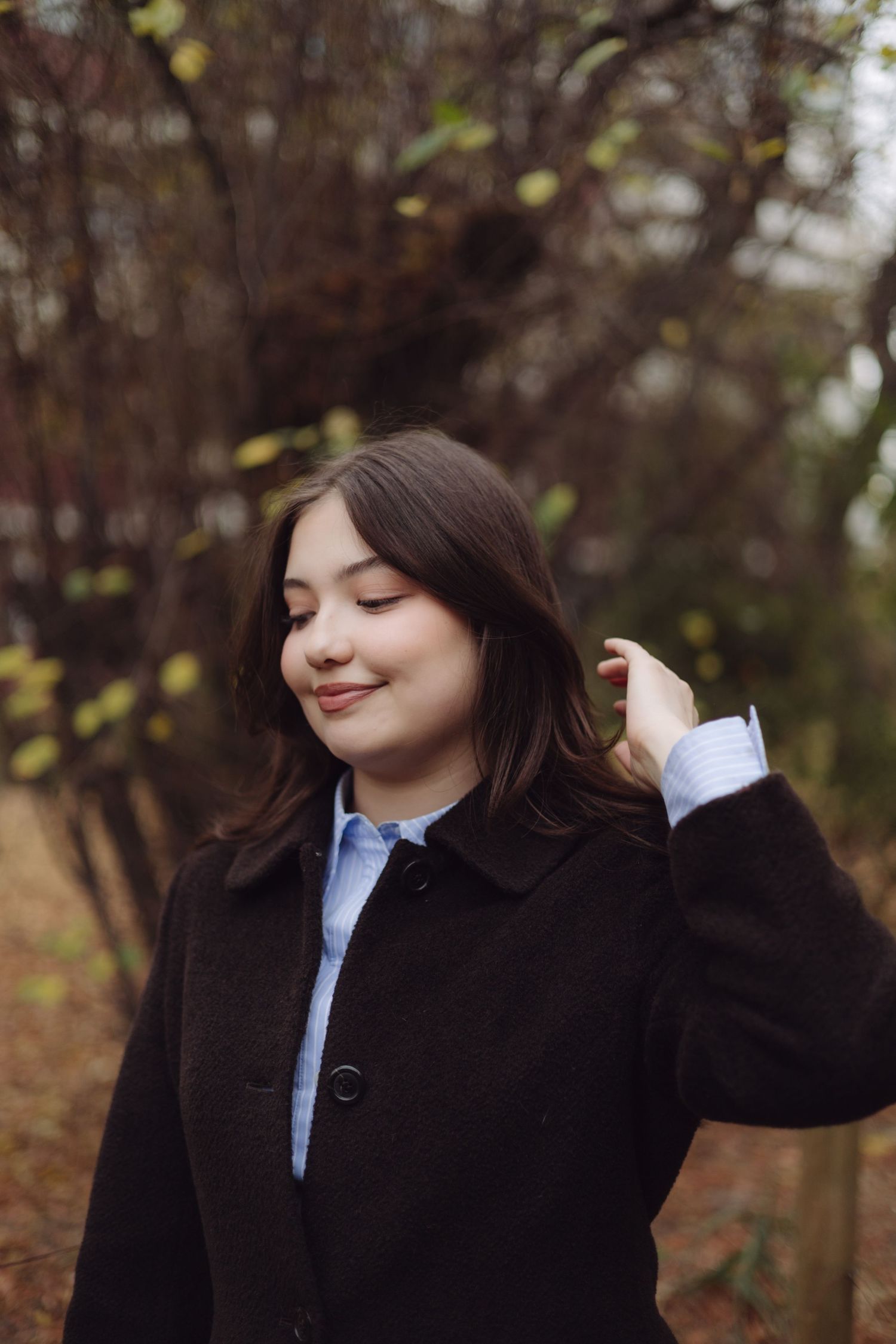 Young woman in dark coat and blue shirt smiling outdoors in autumn park.