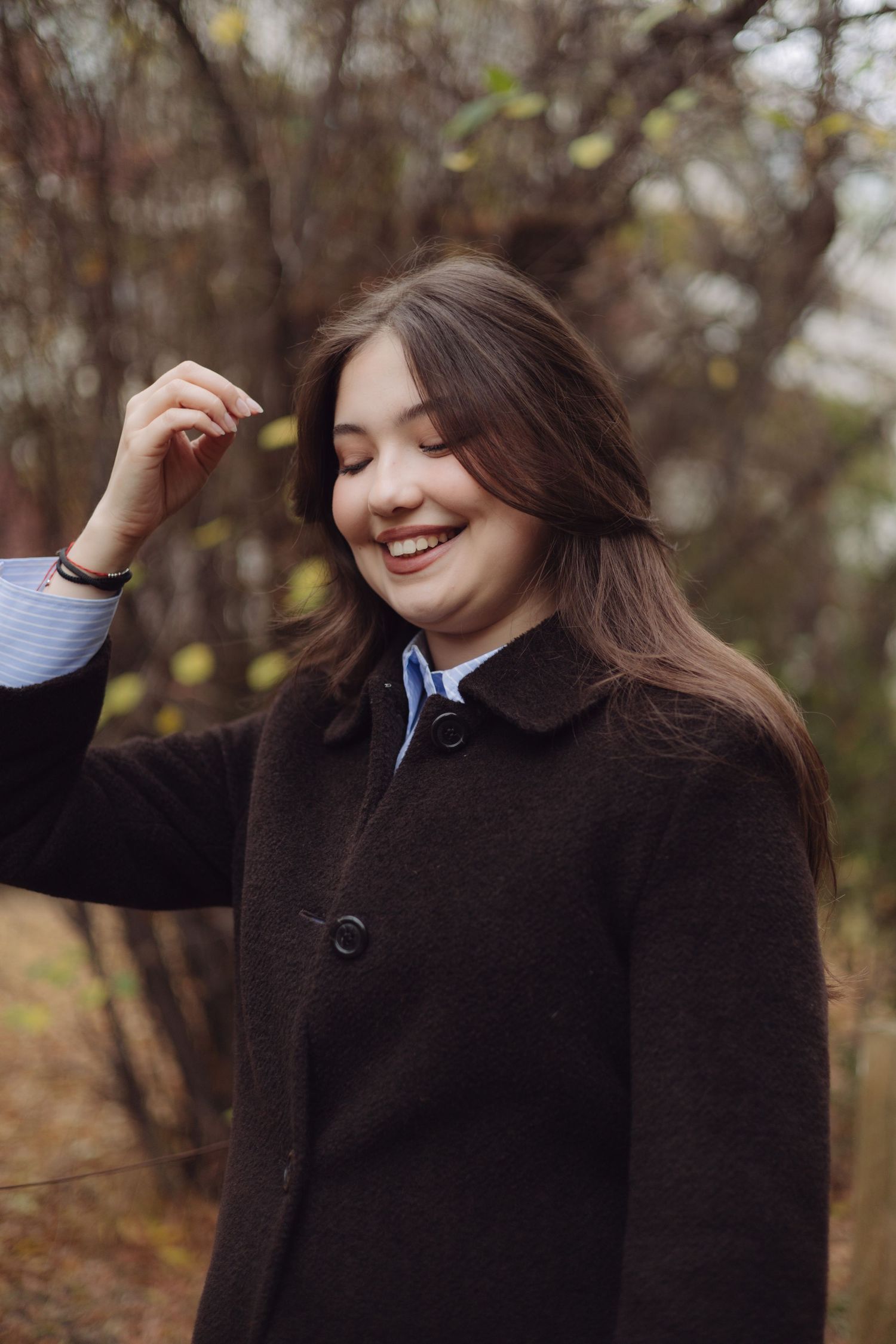 Young woman with long brown hair smiling outdoors in autumn, wearing a dark coat.