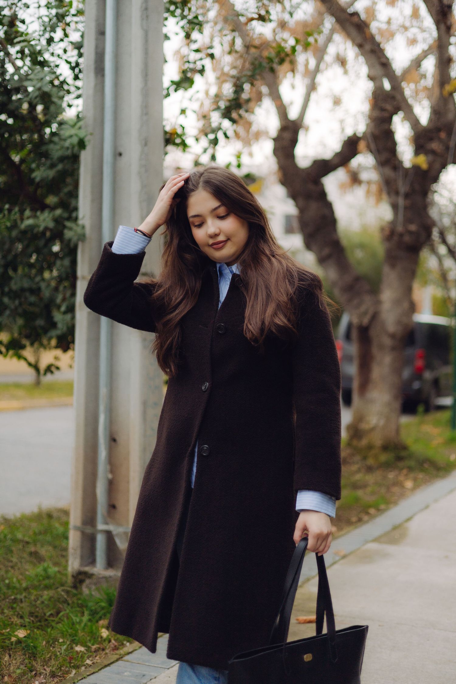 Young woman in dark coat carrying black handbag walks along tree-lined sidewalk, hand in hair, smiling softly.