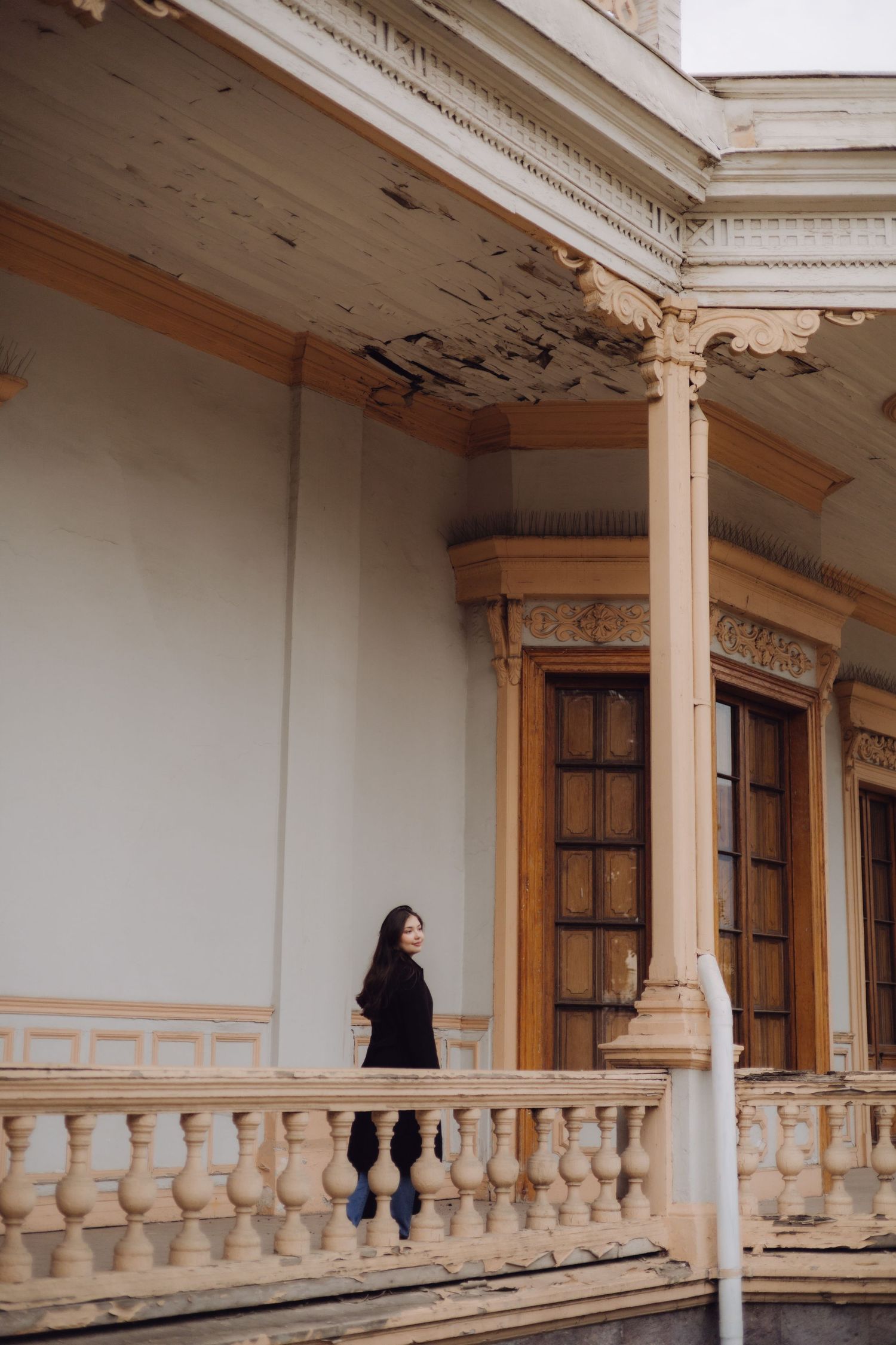 Woman in black outfit standing on ornate balcony of historic building with decorative columns.