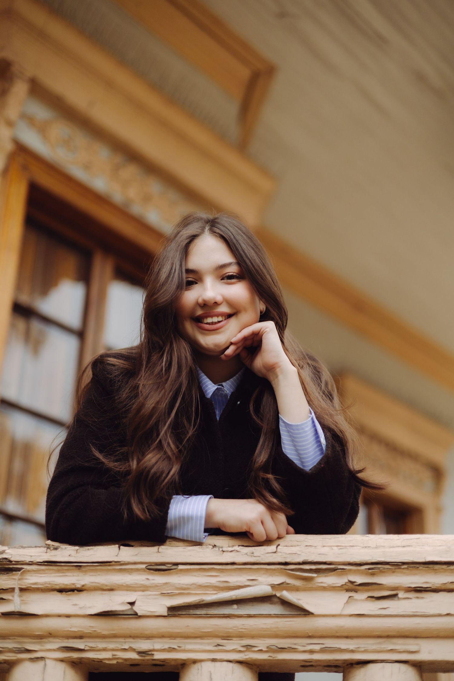 Smiling young woman with long brown hair leans on wooden railing wearing black blazer and blue shirt.
