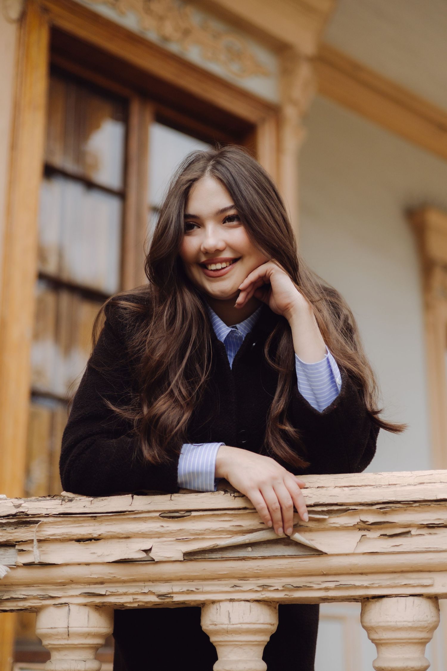 Smiling young woman in dark blazer leaning on wooden balcony railing of vintage building.