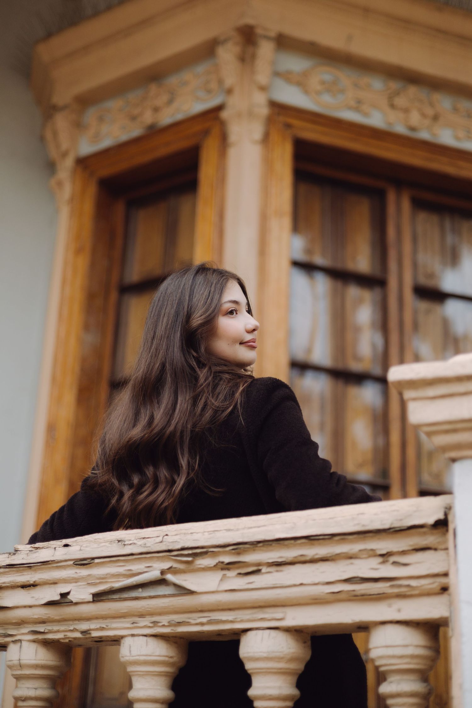Young brunette woman leaning on vintage wooden balcony railing of ornate building.