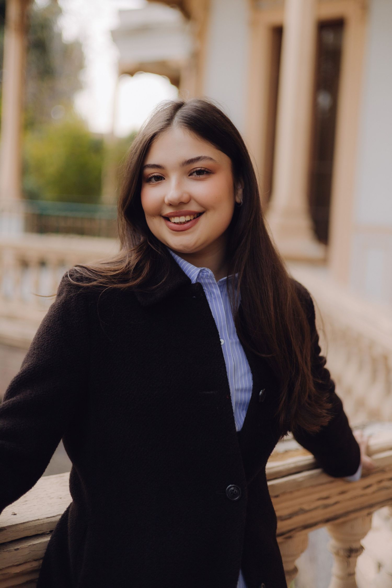 Smiling young woman in black coat and blue shirt leaning on ornate stone balustrade outdoors.