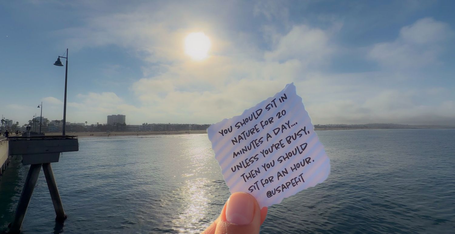 Hand holding a note card with inspirational quote near a sunny ocean pier with blue skies and calm waters.