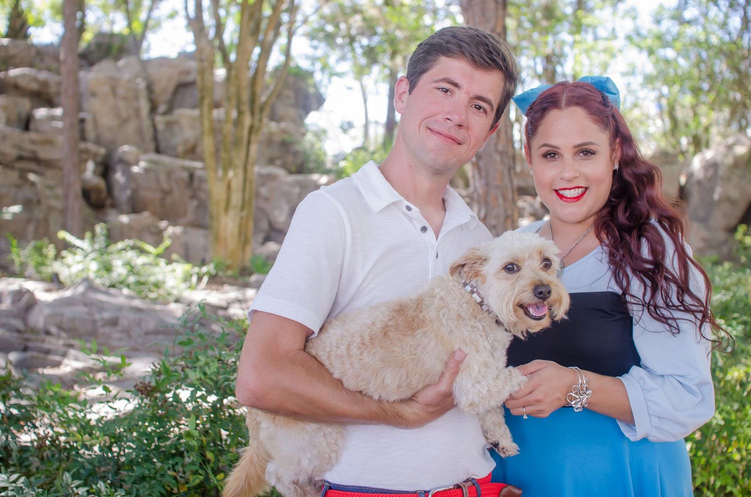 A smiling couple holds a fluffy dog outdoors near rocky scenery, the woman with red hair wearing a blue top.