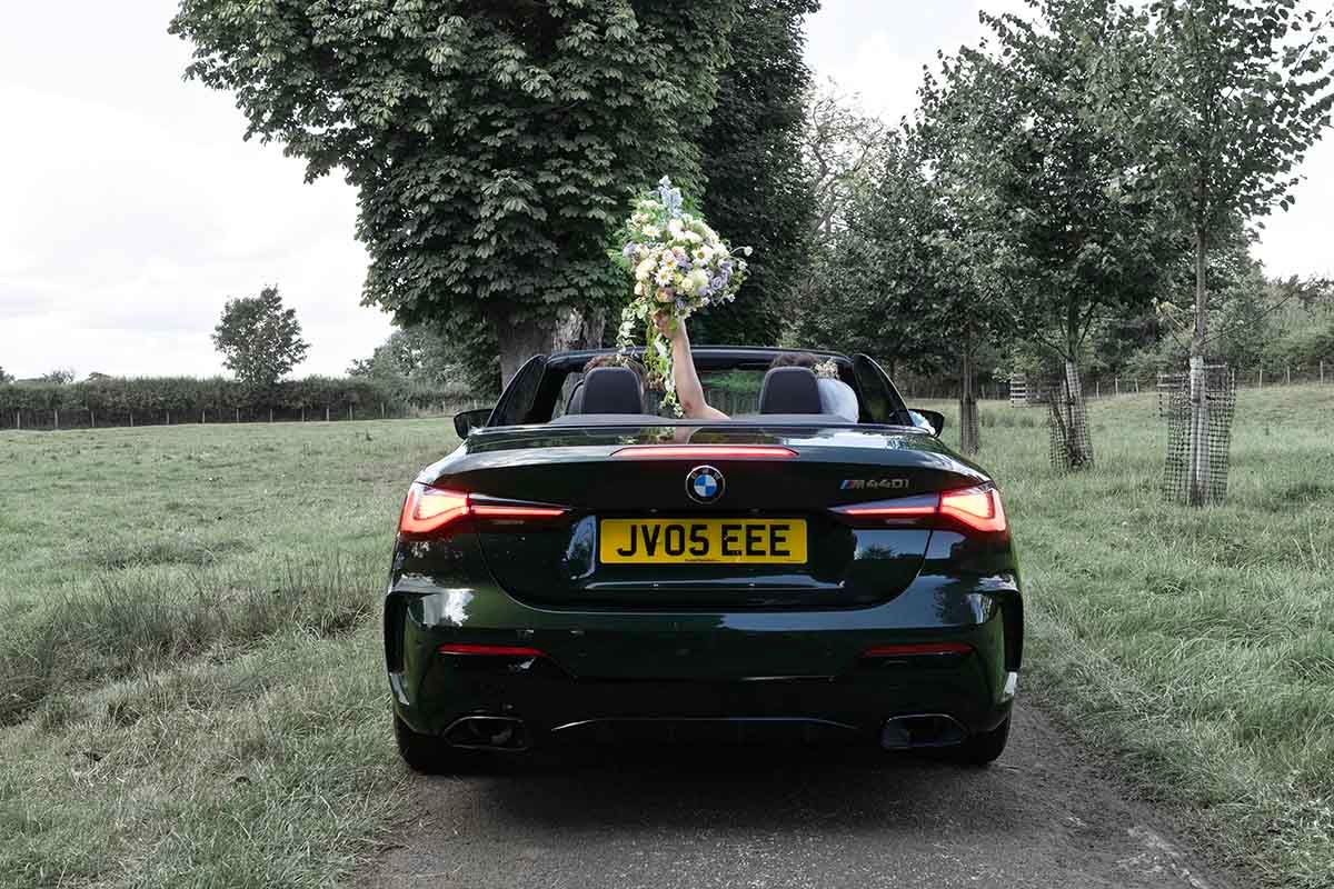 Dark green BMW convertible with bouquet on trunk, parked on rural path, rear view showing the bride