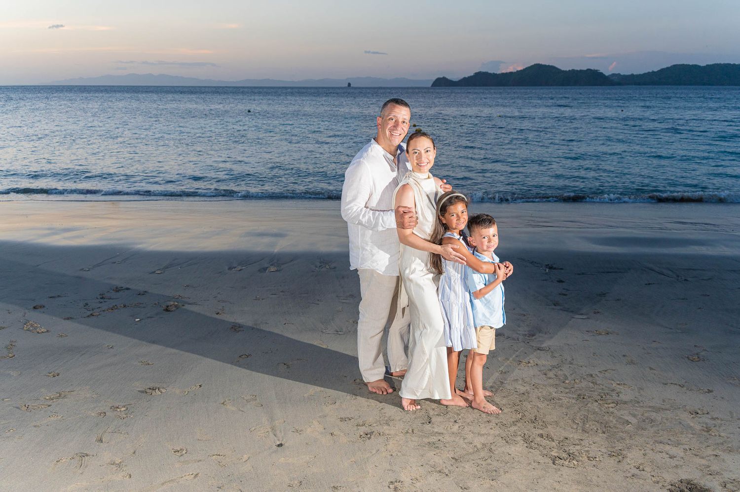 Family of four posing on a sandy beach at sunset, wearing white and blue outfits with ocean waves in the background.