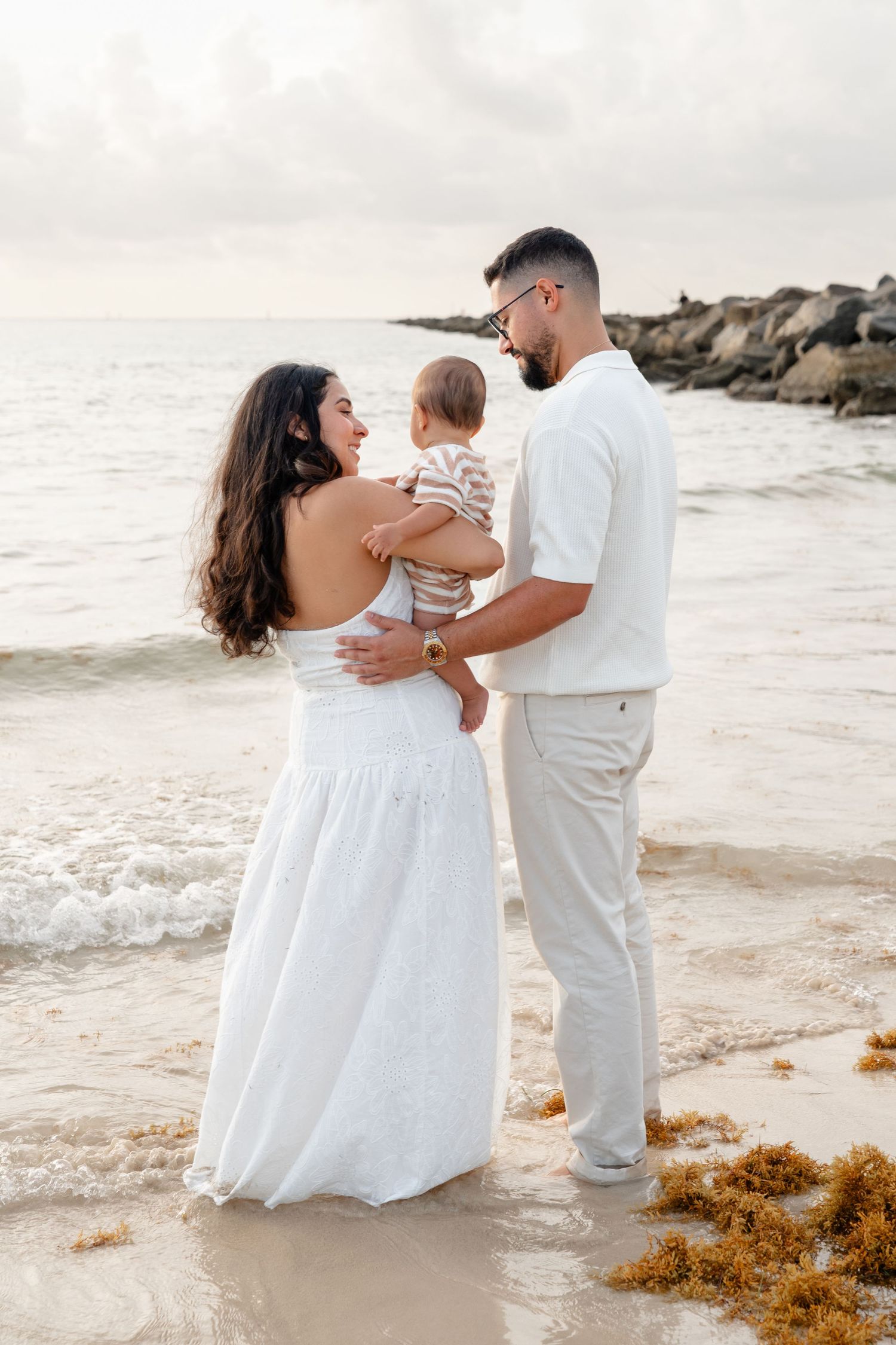 A young family in white outfits stands on a beach at sunset, with the father holding their baby while the mother smiles lovingly.