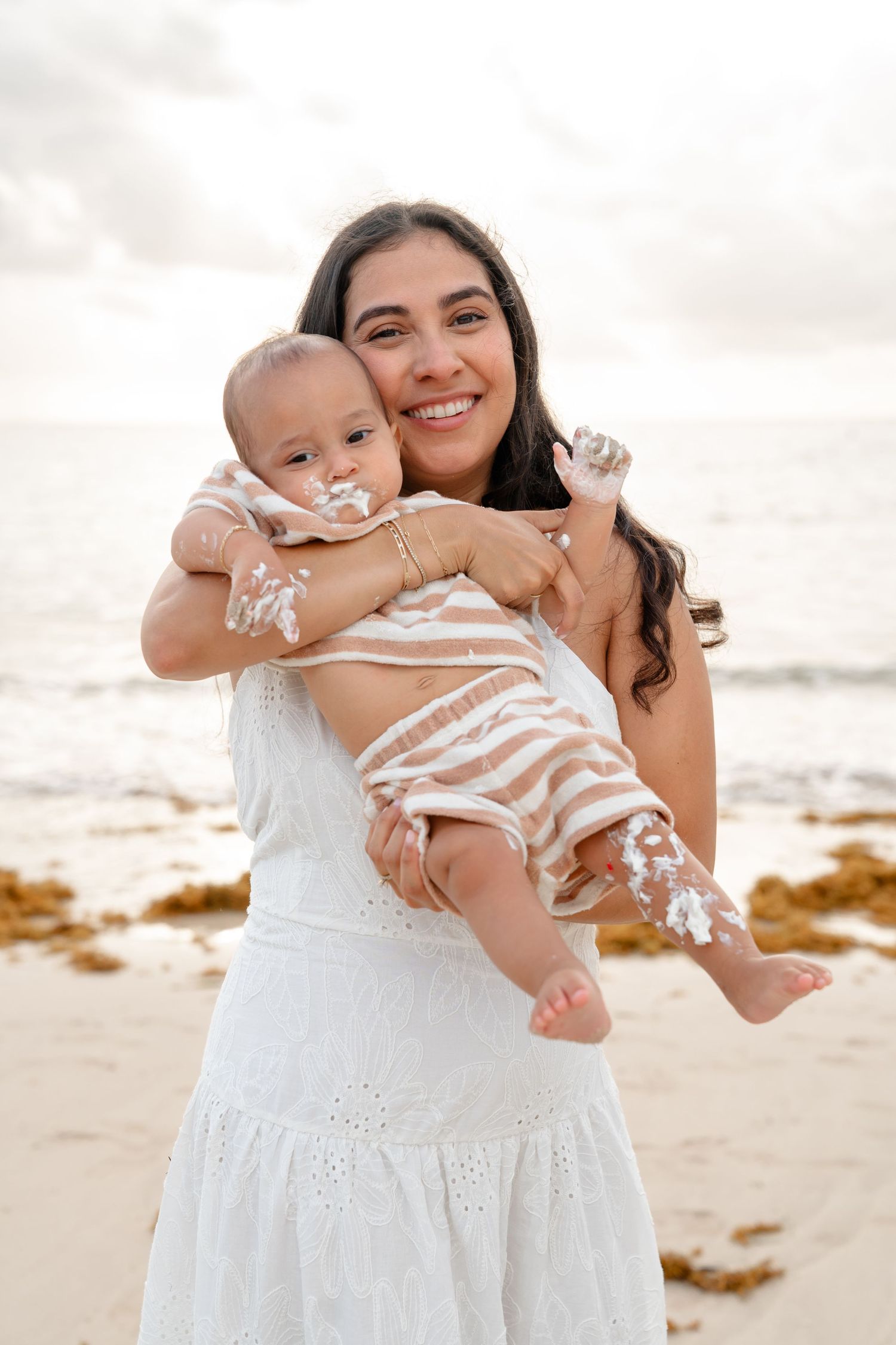 Smiling mother in white dress holds baby with cake on face at beach.