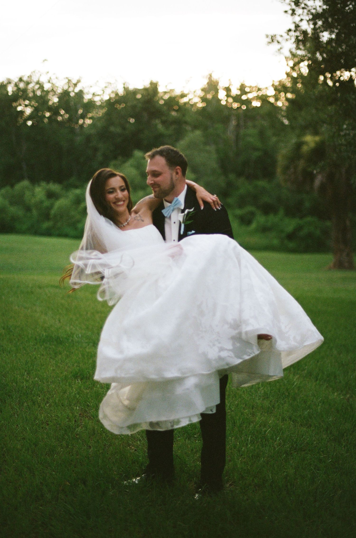 Groom carries bride in white wedding dress and veil on green lawn at sunset.