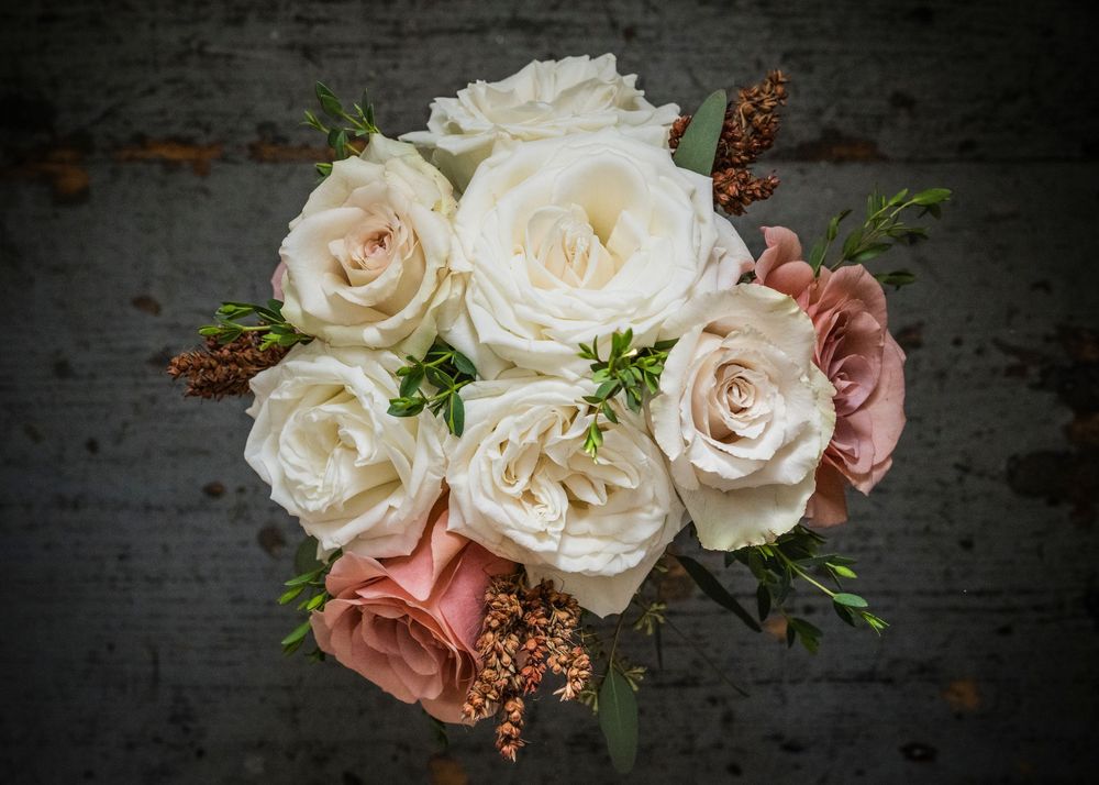Bridal bouquet of cream and blush pink roses with green foliage and rust-colored accents against a weathered dark wood background.