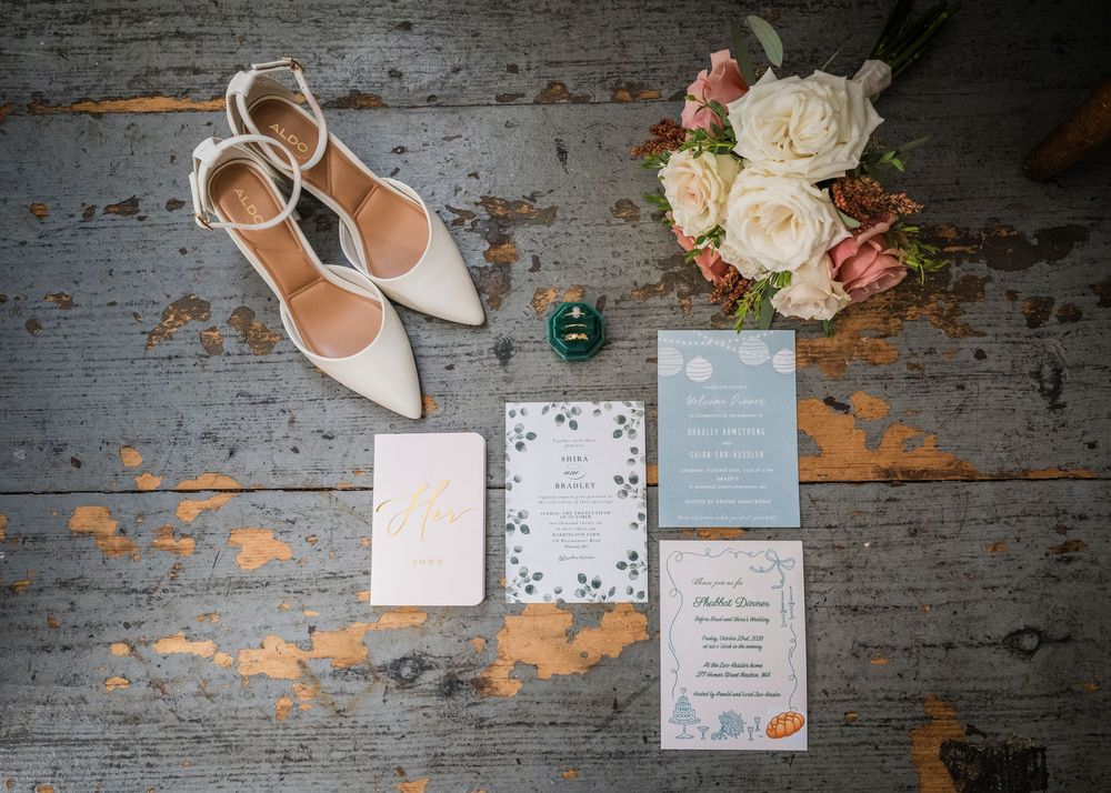 Wedding flat lay with ivory ankle-strap heels, a peach and white bouquet, a ring box, and invitation cards on weathered wood.