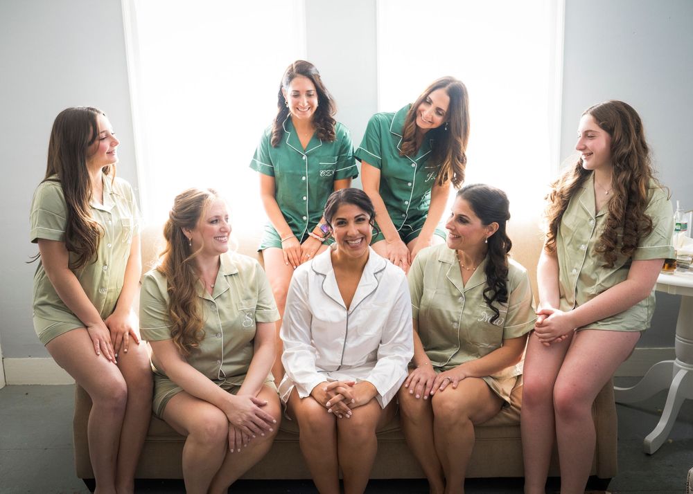 Bride in a white button-up shirt sits among six smiling bridesmaids in matching green pajamas on a couch while getting ready.