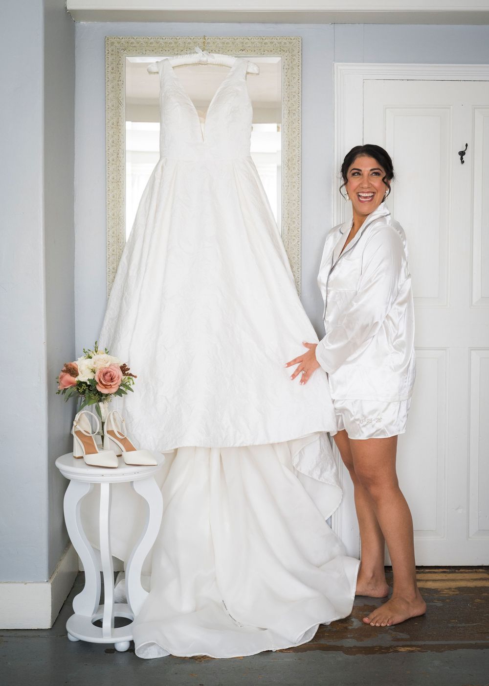 Smiling bride in a satin robe stands barefoot beside her wedding dress hanging on a mirror, with heels and a bouquet nearby.