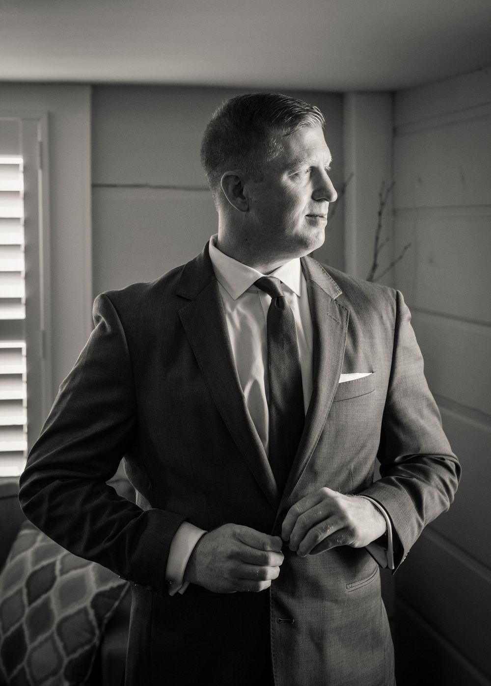 Black and white portrait of a groom in a suit adjusting his jacket button while gazing out a window.