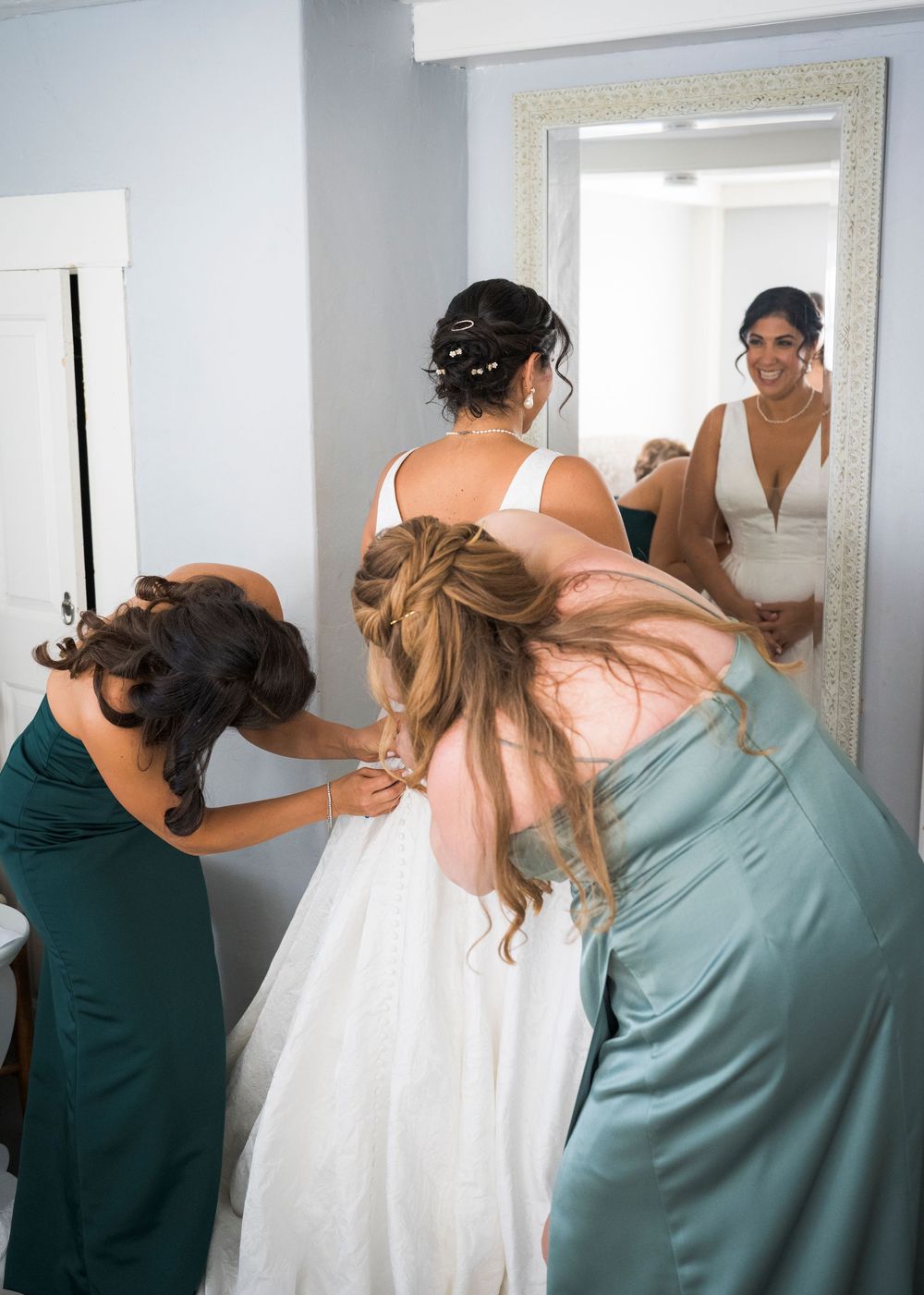 Two bridesmaids in green dresses help button the bride's wedding gown while she smiles at her reflection in a large mirror.