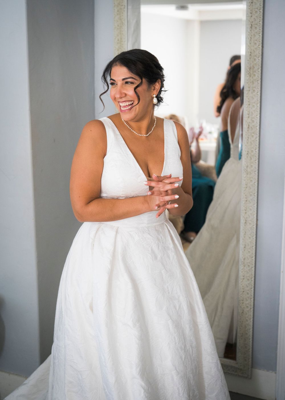 A smiling bride in an elegant white V-neck wedding gown clasps her hands beside a tall mirror while getting ready for her wedding.