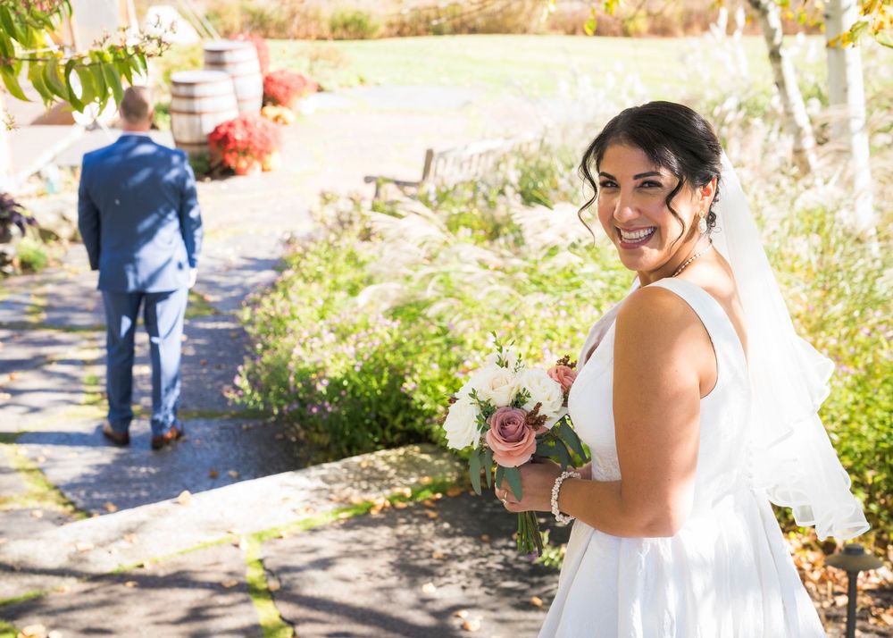 Smiling bride in a white gown and veil holds a white and pink bouquet on a garden path while a groom in a blue suit walks away.