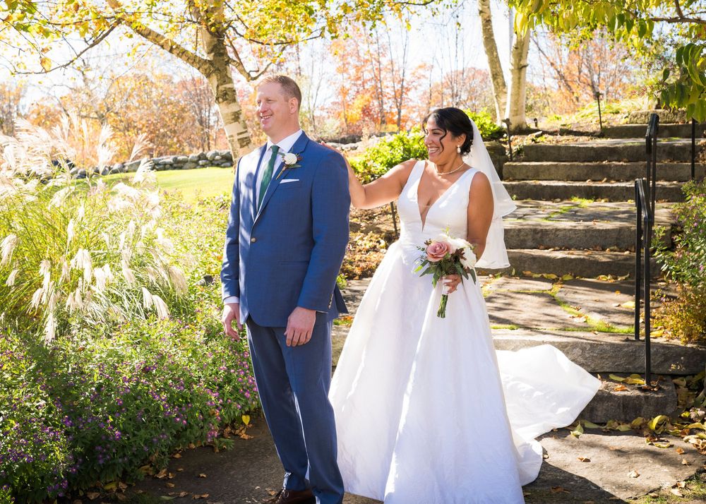 Groom in a navy suit and bride holding a pink bouquet share a happy moment near stone steps surrounded by colorful autumn foliage.