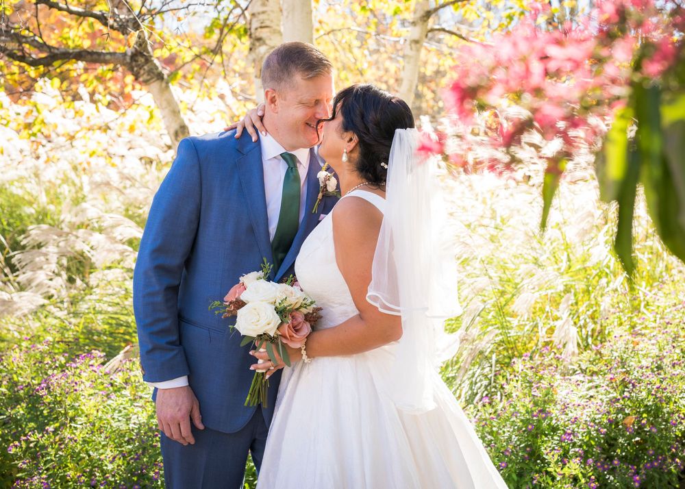 Bride in a white gown and veil kisses groom in a blue suit while holding a bouquet among colorful autumn foliage.