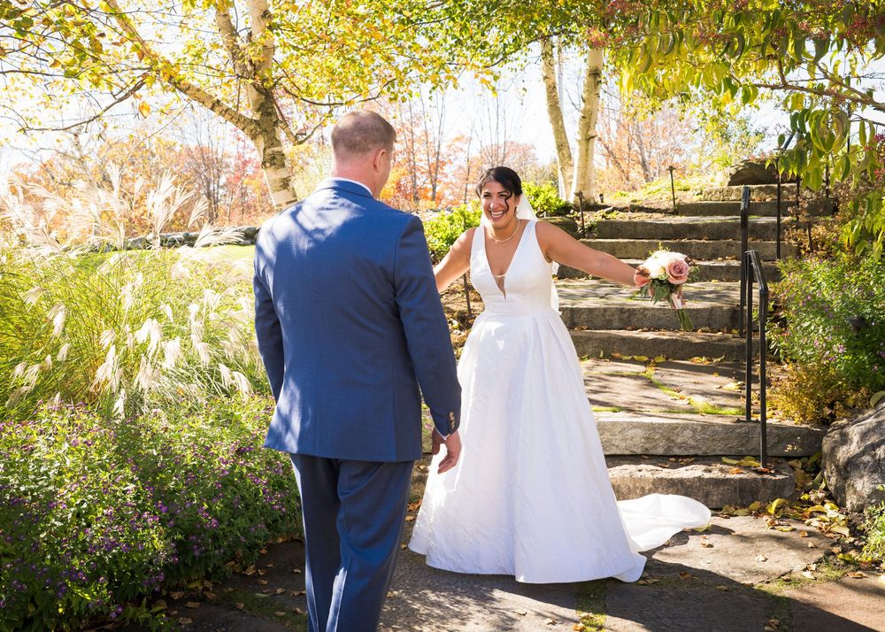 Bride in a white gown smiles at the groom in a navy suit as they meet on stone steps surrounded by colorful autumn foliage.