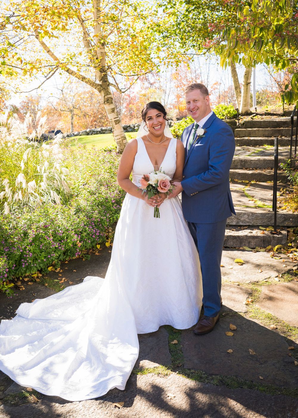 Smiling bride in a white gown and groom in a blue suit pose with a bouquet on a stone path surrounded by autumn foliage.