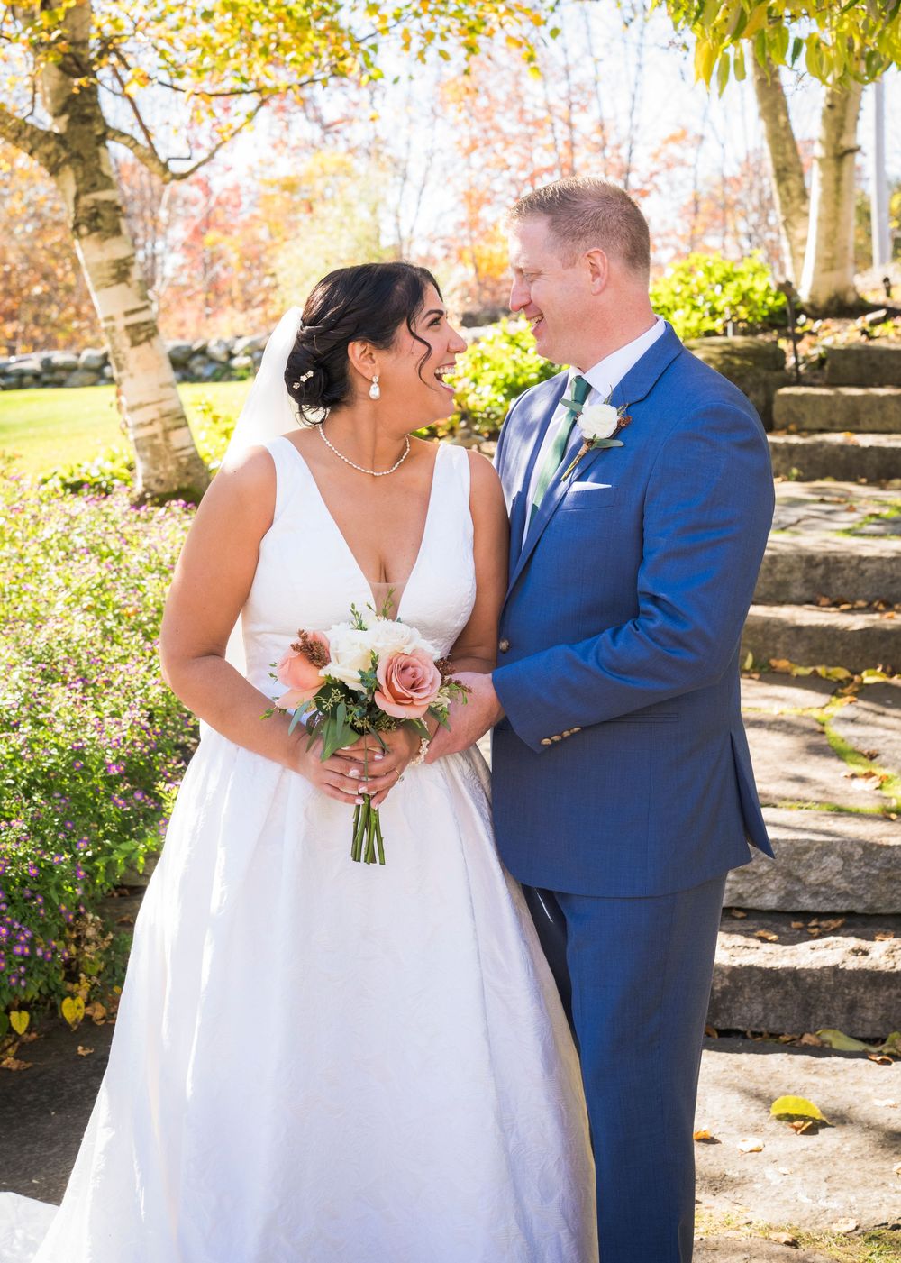 Smiling bride in a white gown holding a rose bouquet laughs with her groom in a blue suit on stone steps surrounded by autumn foliage.