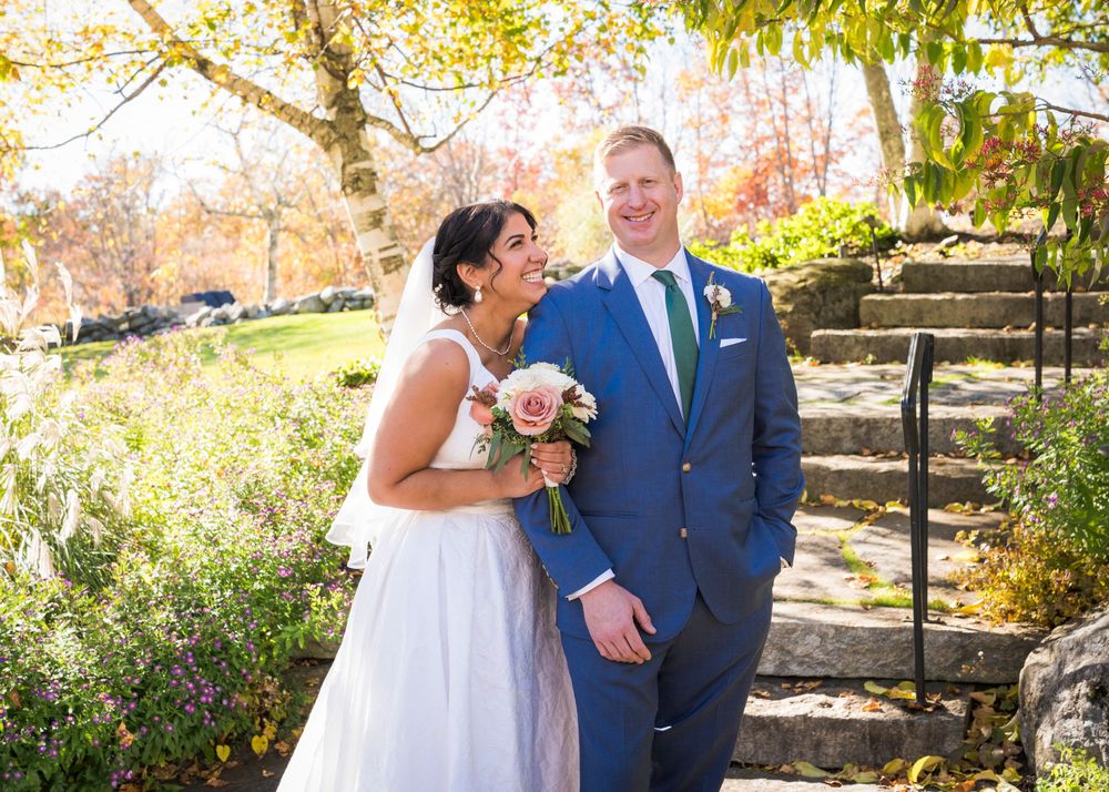 Happy bride and groom in a blue suit share a joyful moment beside stone steps with colorful fall trees and a pink and white bouquet.