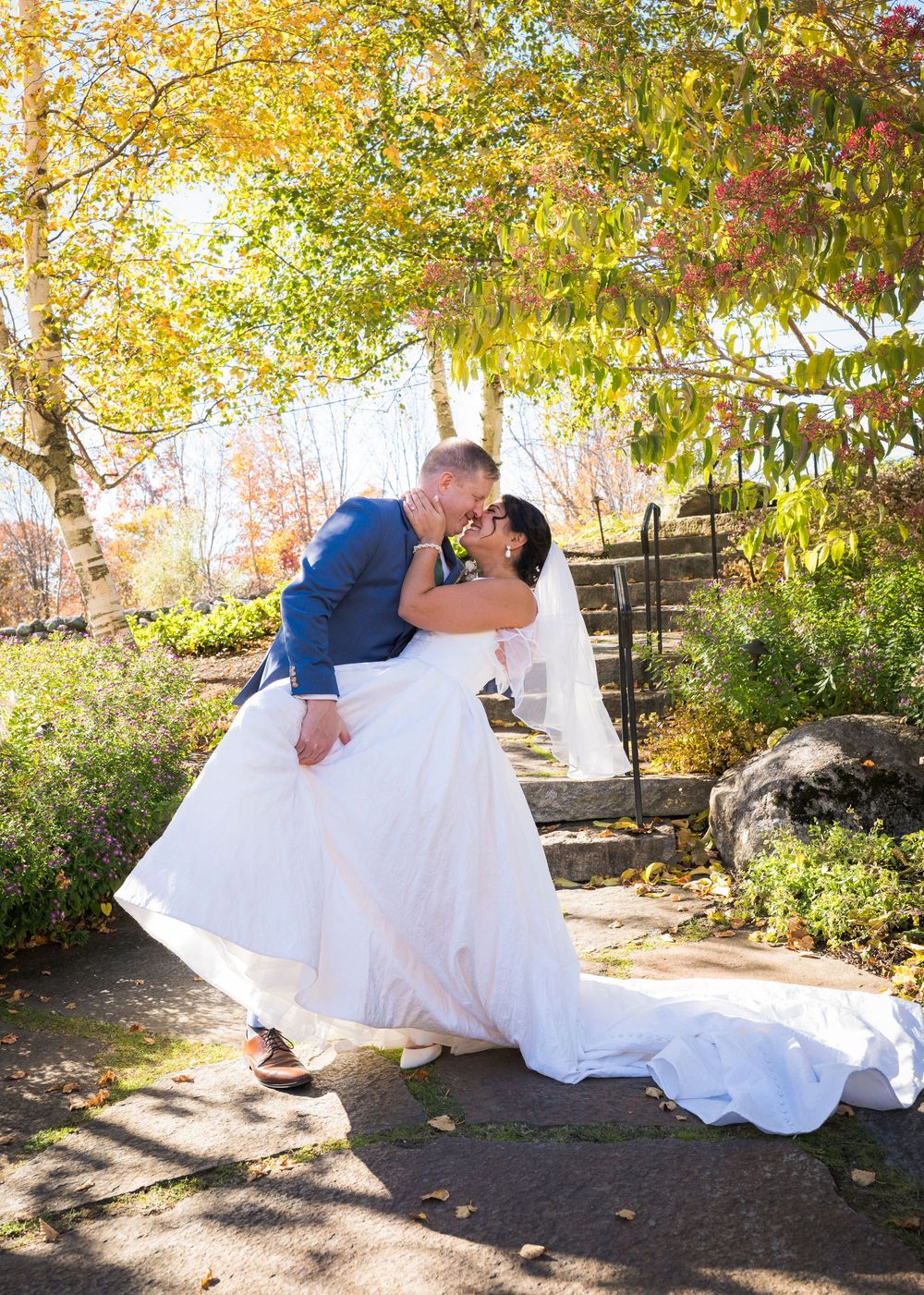Groom in a blue jacket dips and kisses the bride in her flowing white gown on a stone path surrounded by colorful autumn foliage.