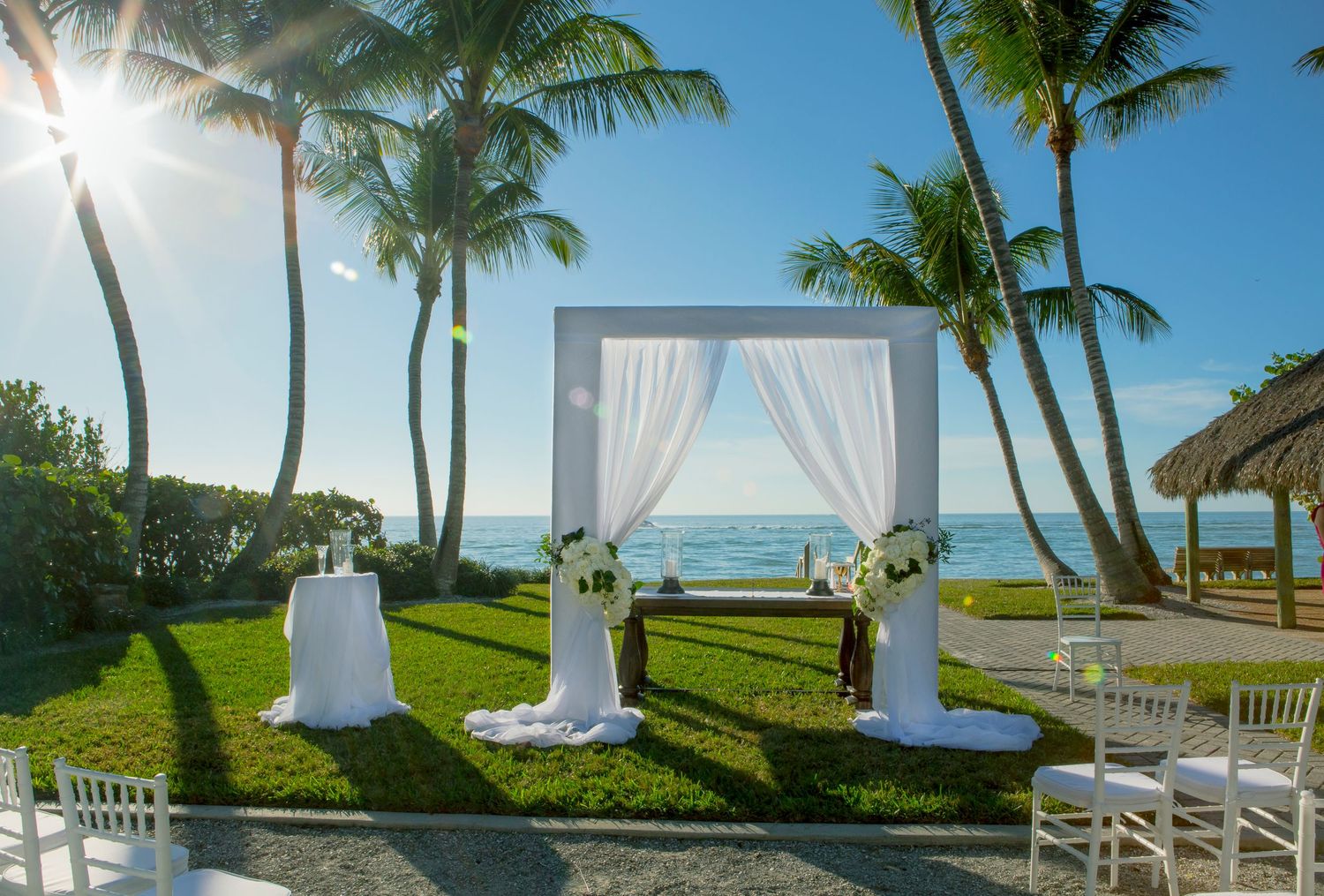 Elegant outdoor beach wedding ceremony setup with white draped arch, palm trees, and ocean view.