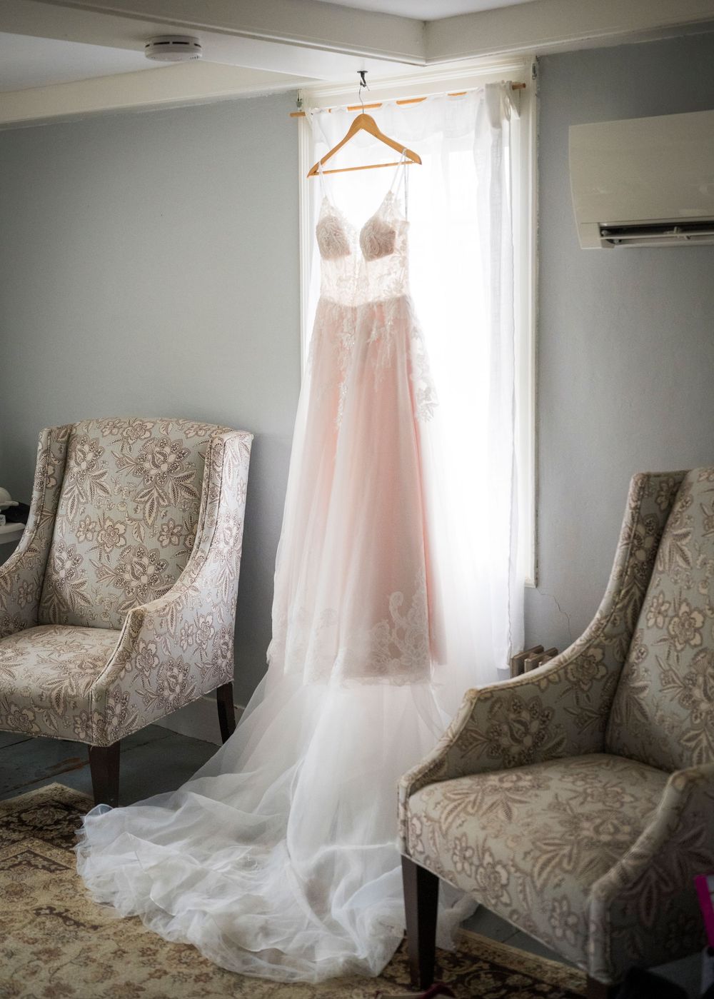 Elegant blush pink lace wedding gown hanging on a wooden hanger in a softly lit bridal suite.