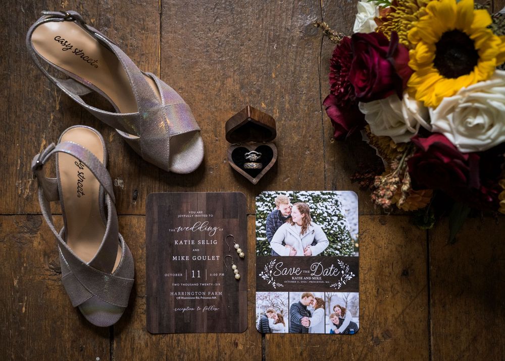 Fall wedding flat lay with bridal shoes, ring box, bouquet, and save the date cards on rustic wood floor.