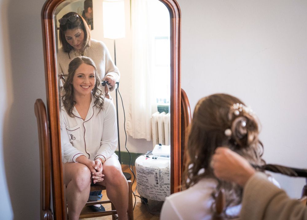 Bride smiles in mirror reflection while bridesmaid styles her hair for wedding.