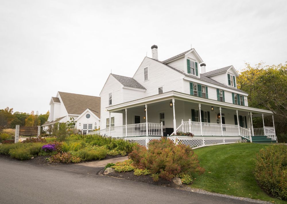 White farmhouse wedding venue with wrap-around porch and green shutters surrounded by lush gardens.