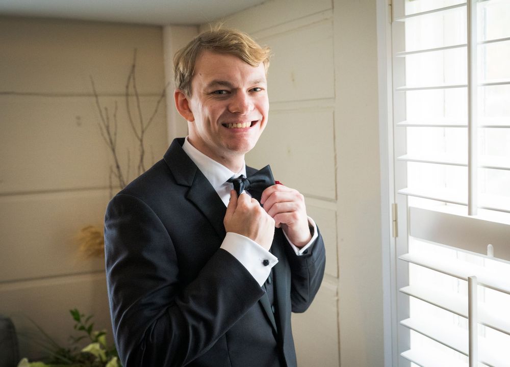 Young groom in black tuxedo adjusting bow tie near window, smiling on wedding day.