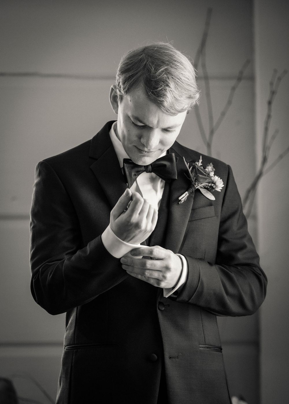 Dapper groom in black tuxedo adjusts cufflinks with floral boutonniere, black and white.
