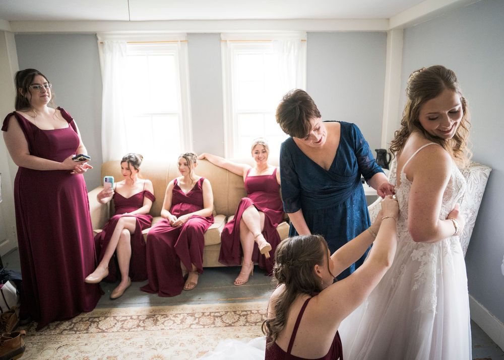 Bridesmaids in burgundy dresses helping bride into her white wedding gown before the ceremony.