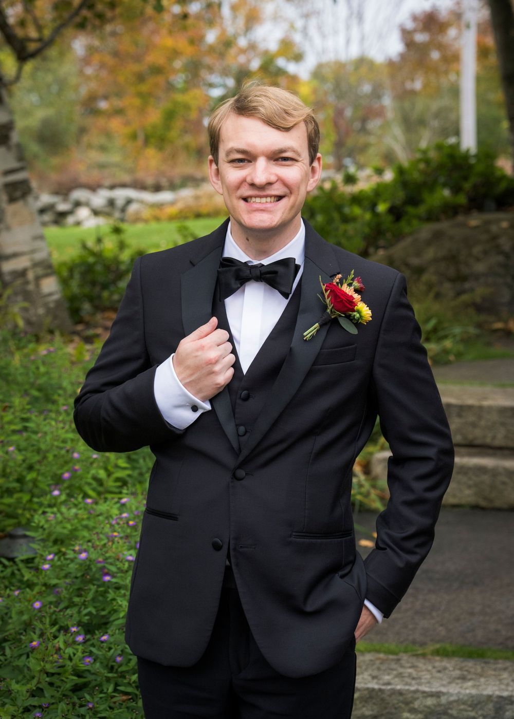 Smiling groom in black tuxedo with colorful boutonniere poses outdoors in autumn garden.