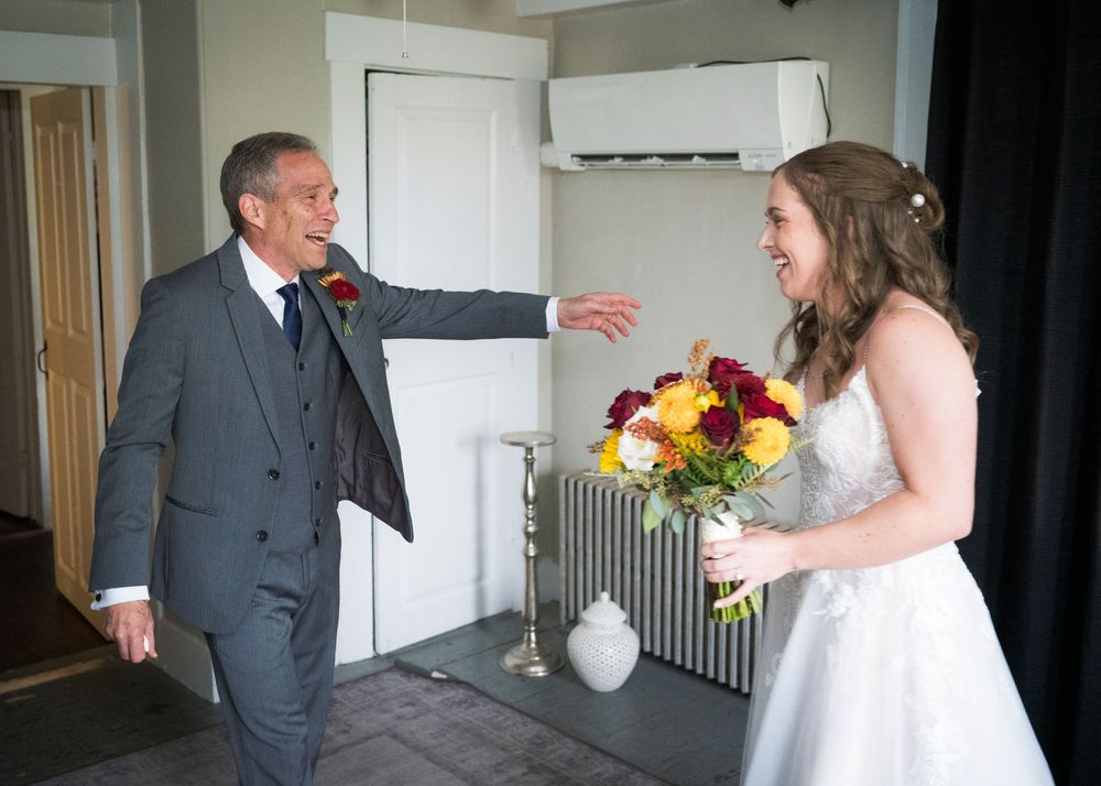 Father reacts with joy seeing bride in wedding dress holding colorful bouquet indoors.