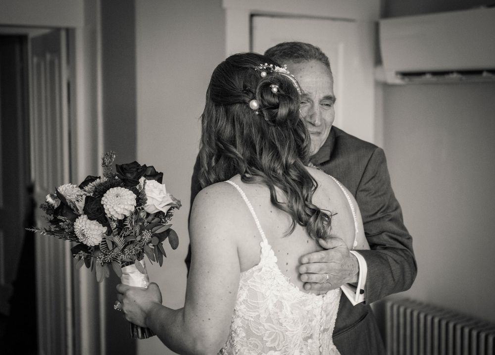 Black and white photo of father embracing bride holding bouquet before wedding ceremony.