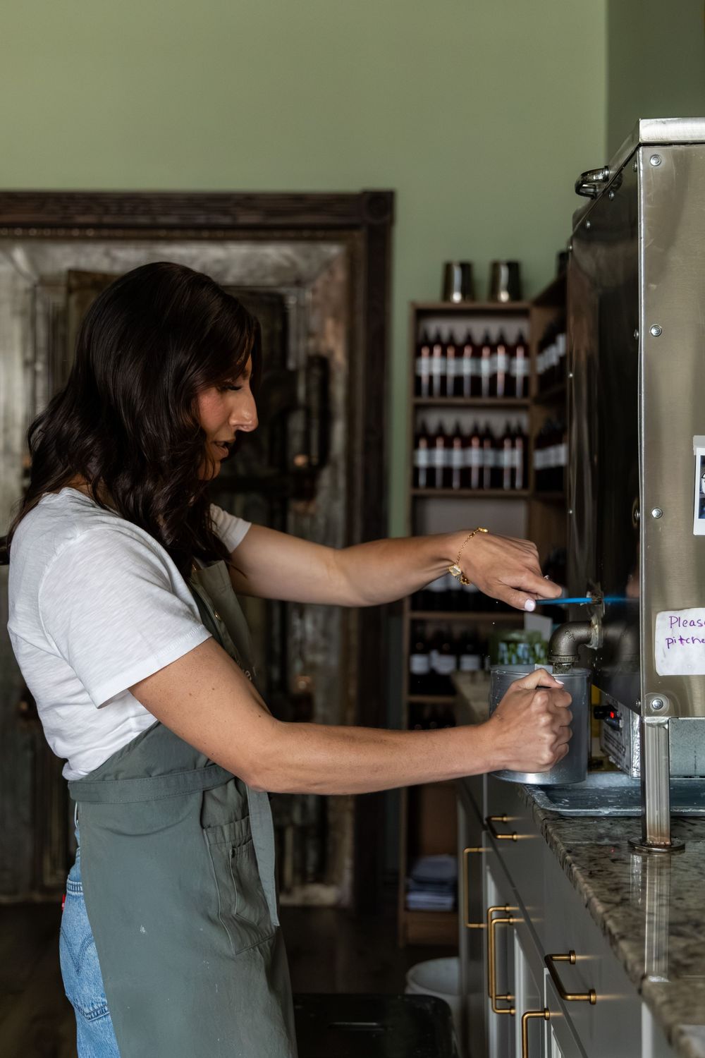 Woman in apron filling a metal container at a dispenser in a shop with shelved bottles in background.
